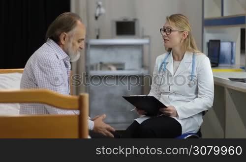 Friendly woman doctor with clipboard and in spectacles talking with senior male patient at hospital office. Positive female physician with stethoscope in lab coat explaining good medical results and giving treatment advice to happy elderly man.
