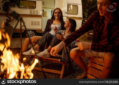 Friends cooking on campfire, picnic at camping in the forest. Youth having summer adventure on rv, camping-car on background. Two couples leisures, travelling with trailer. Friends cooking on campfire, picnic at camping