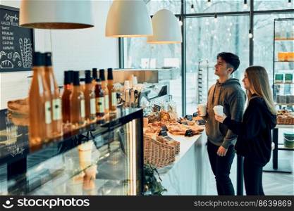 Friends doing shopping in a coffee shop. Young man and woman having chat while picking out the pastry, bakery’s goods and hot drinks standing at counter in a coffee shop. People buying coffee and sweet snacks to go