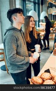 Friends doing shopping in a coffee shop. Young man and woman having chat while picking out the pastry, bakery’s goods and hot drinks standing at counter in a coffee shop. People buying coffee and sweet snacks to go