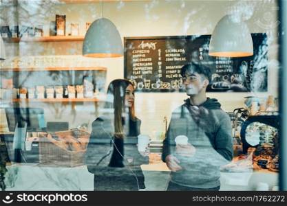 Friends having a chat, holding coffee cups, standing in a coffee shop and waiting for the their orders. Man and woman having a break drinking coffee in cafe. Interior view through the window