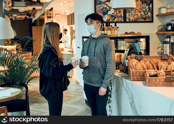 Friends having a chat, holding coffee cups, standing in a coffee shop and waiting for the orders. People wearing the face masks to avoid virus infection and to prevent the spread of disease in time of coronavirus