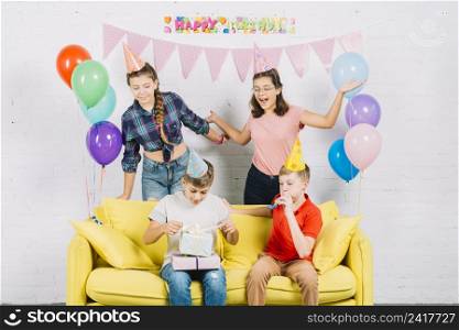 friends having fun while boy sitting sofa unwrapping birthday gifts home