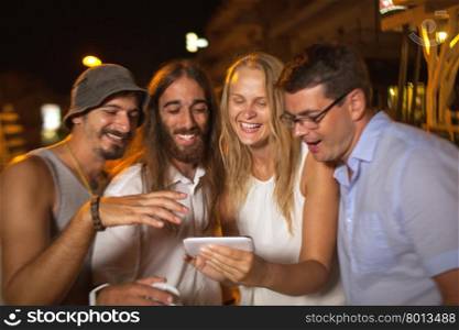 Friends looking at great shots of today evening. Young woman and three men looking through the photos on mobile phone in the street at night. Happy faces showing they got great and funny shots