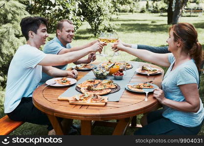 Friends making toast during summer picnic outdoor dinner in a home garden. Close up of people holding wine glasses with white wine over table with pizza, salads and fruits. Dinner in a orchard in a backyard. Friends making toast during summer picnic outdoor dinner in a home garden