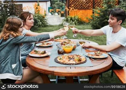 Friends making toast during summer picnic outdoor dinner in a home garden. Close up of people holding wine glasses with white wine over table with pizza, salads and fruits. Dinner in a orchard in a backyard. Friends making toast during summer picnic outdoor dinner in a home garden