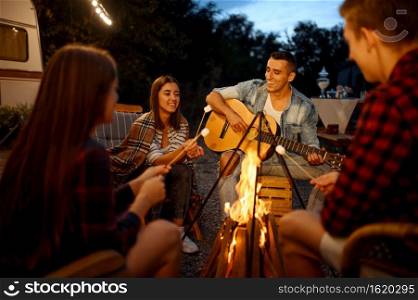 Friends playing on guitar by the bonfire, picnic at c&ing in the forest. Youth having summer adventure on rv, c&ing-car on background. Two couples leisures, travelling with trailer. Friends playing on guitar by the bonfire, c&ing