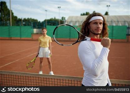 Friends playing tennis on court. Focus on confident man player holding racket on shoulder and looking in camera. Weekend sports recreation for young people couple. Couple of friends playing tennis on court with focus on confident man