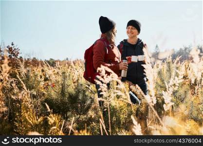 Friends relaxing and enjoying the coffee during vacation trip. People standing on trail drinking coffee. Couple with backpacks hiking through tall grass along path in mountains. Active leisure time close to nature