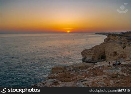 Friends seated on cliff and watching Sunset at Atlantic ocean coast, in Carvoeiro beach clifs, Algarve, Portugal