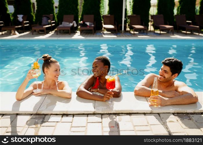 Friends with beverages pose at the edge of the pool. Happy people having fun on summer vacations, holiday party at the poolside outdoors. One man and two women are sunbathing. Friends with beverages pose at the edge of pool