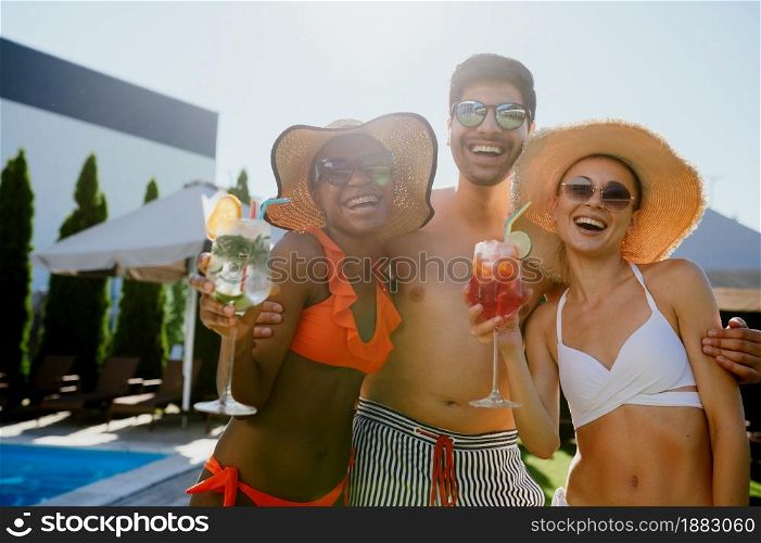 Friends with coctails pose at the pool in hotel. Happy people having fun on summer vacations, holiday party at the poolside outdoors. One man and two women are sunbathing. Friends with coctails pose at the pool in hotel