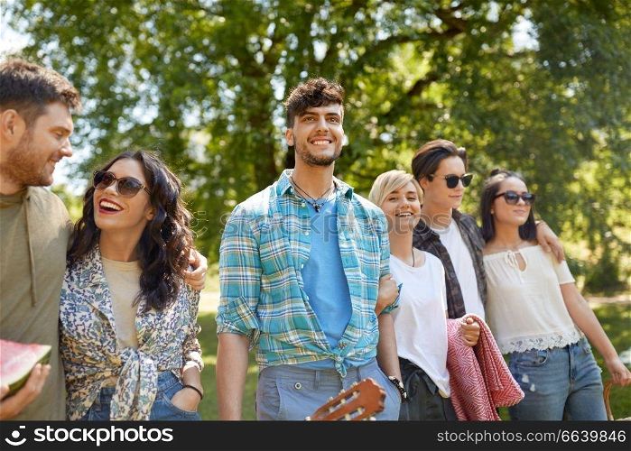 friendship and leisure concept - group of happy friends with guitar and blanket going to picnic at summer park. friends with guitar going to picnic at park