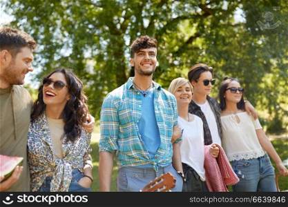 friendship and leisure concept - group of happy friends with guitar and blanket going to picnic at summer park. friends with guitar going to picnic at park
