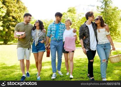 friendship and leisure concept - group of happy friends with guitar, picnic basket, blanket and watermelon at summer park. friends with guitar going to picnic at park
