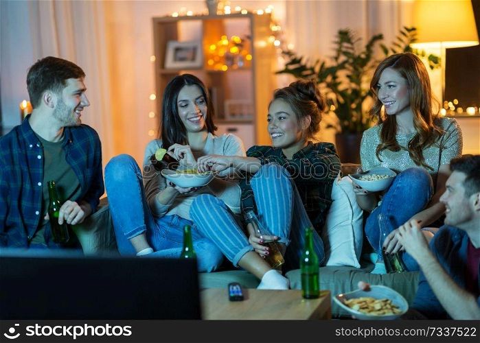 friendship, and leisure concept - happy friends with non-alcoholic drinks and snacks watching tv at home in evening. friends with drinks and snacks watching tv at home