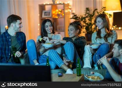 friendship, and leisure concept - happy friends with non-alcoholic drinks and snacks watching tv at home in evening. friends with drinks and snacks watching tv at home
