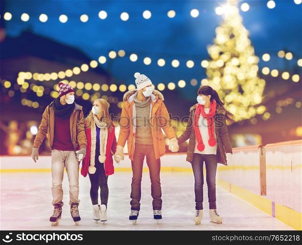 friendship, christmas and leisure concept - friends wearing face protective medical masks for protection from virus disease holding hands at outdoor skating rink over holiday lights background. friends in masks on christmas skating rink