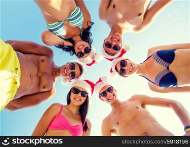 friendship, christmas, summer vacation, holidays and people concept - group of smiling friends wearing swimwear and santa helper hats standing in circle over blue sky