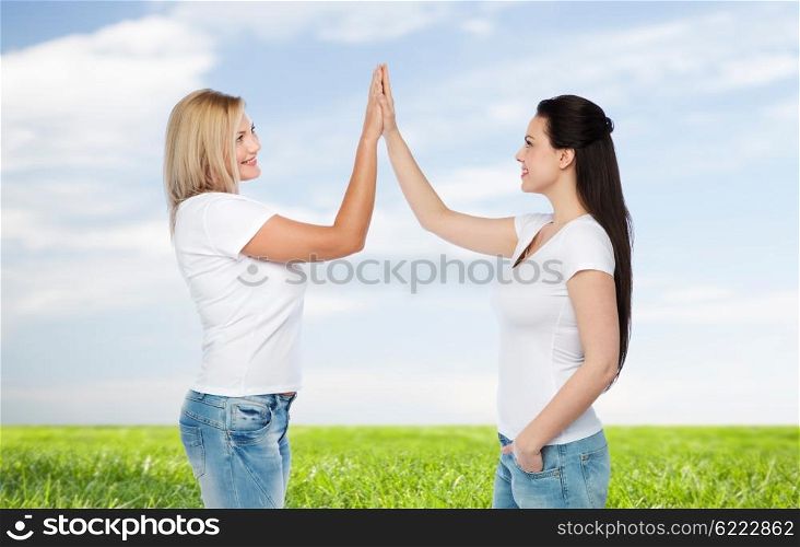 friendship, diverse, body positive, gesture and people concept - group of happy different women in white t-shirts making high five over blue sky and grass background