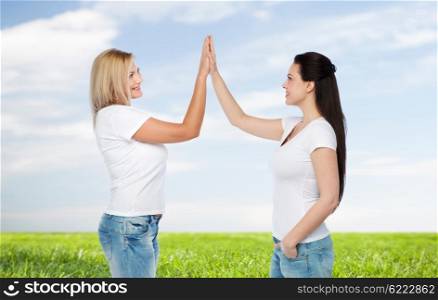 friendship, diverse, body positive, gesture and people concept - group of happy different women in white t-shirts making high five over blue sky and grass background