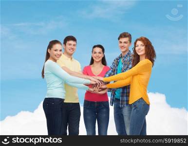 friendship, dream, teamwork, gesture and people concept - group of smiling teenagers with hands on top of each other over blue sky with white cloud background
