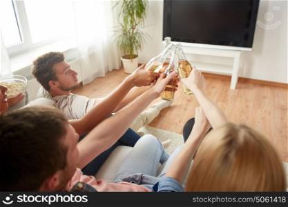 friendship, drink, alcohol, holidays and people concept - happy friends clinking beer bottles and watching tv at home. friends clinking beer and watching tv at home