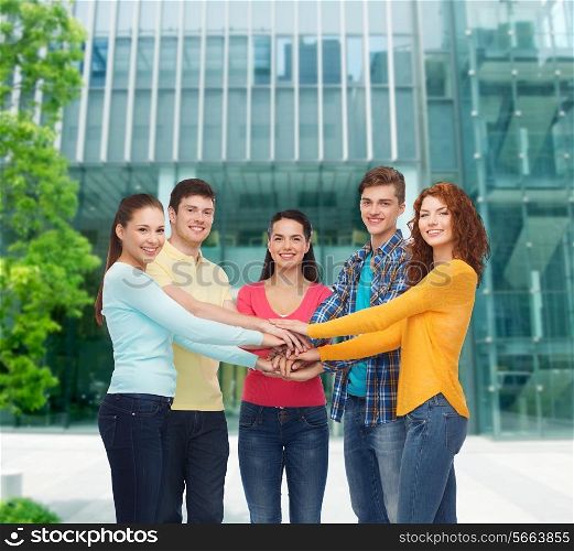 friendship, education, business, gesture and people concept - group of smiling teenagers with hands on top of each other over campus background