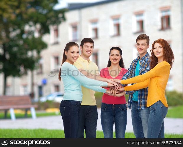 friendship, education, teamwork, gesture and people concept - group of smiling teenagers putting hand on top of each other over campus background