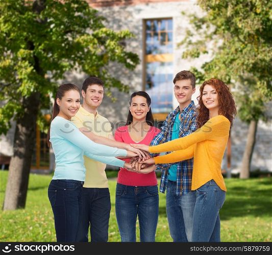 friendship, education, teamwork, gesture and people concept - group of smiling teenagers putting hand on top of each other over campus background