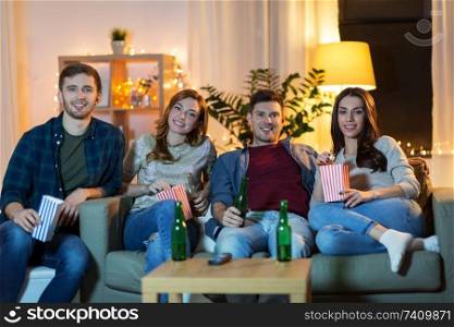 friendship, entertainment and leisure concept - happy friends with non-alcoholic drinks and popcorn watching tv at home in evening. friends with beer and popcorn watching tv at home