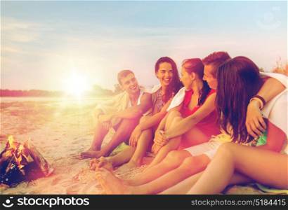 friendship, happiness, summer vacation, holidays and people concept - group of smiling friends sitting near fire on beach. smiling friends in sunglasses on summer beach