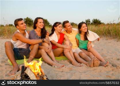 friendship, happiness, summer vacation, holidays and people concept - group of smiling friends sitting near fire on beach