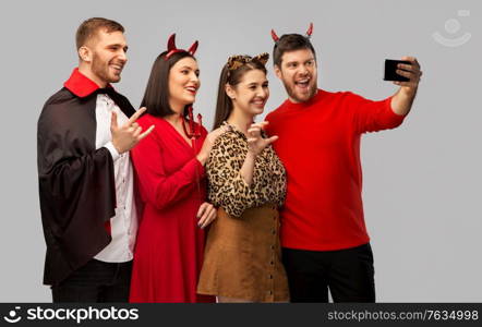 friendship, holiday and people concept - group of happy smiling friends in halloween costumes of vampire, devil and cheetah taking selfie by smartphone over grey background. happy friends in halloween costumes taking selfie