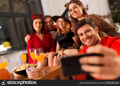 friendship, holiday and people concept - group of happy smiling friends in halloween costumes of vampire, devil, witch and cheetah taking selfie by smartphone at home party at night. happy friends in halloween costumes taking selfie