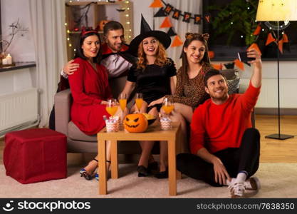 friendship, holiday and people concept - group of happy smiling friends in halloween costumes of vampire, devil, witch and cheetah taking selfie by smartphone at home party at night. happy friends in halloween costumes taking selfie