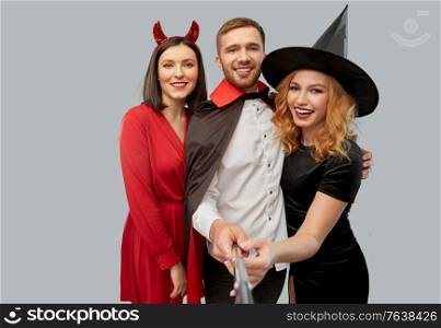 friendship, holiday and people concept - group of happy smiling friends in halloween costumes of witch, devil and vampire taking picture by selfie stick over grey background. happy friends in halloween costumes taking selfie