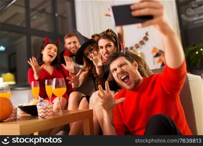 friendship, holiday and people concept - group of happy smiling friends in halloween costumes of vampire, devil, witch and cheetah taking selfie by smartphone at home party at night. happy friends in halloween costumes taking selfie