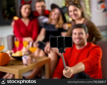 friendship, holiday and people concept - group of happy smiling friends in halloween costumes taking picture with smartphone on selfie stick at home party at night. happy friends in halloween costumes taking selfie