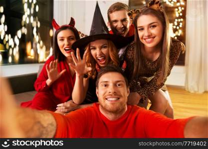 friendship, holiday and people concept - group of happy smiling friends in halloween costumes of vampire, devil, witch and cheetah taking selfie at home party at night. happy friends in halloween costumes taking selfie