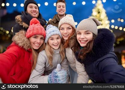 friendship, holidays and people concept - group of happy friends taking selfie outdoors and showing thumbs up gesture over christmas lights background. happy friends taking selfie outdoors at christmas