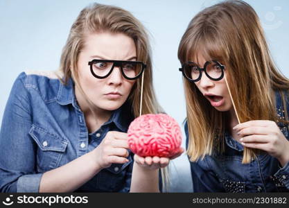 Friendship, human relations concept. Two crazy women friends or sisters wearing jeans shirts and eyeglasses on stick, thinking about solving problem holding fake brain. Two crazy women friends thinking