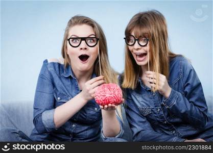 Friendship, human relations concept. Two crazy women friends or sisters wearing jeans shirts and eyeglasses on stick, thinking about solving problem holding fake brain. Two crazy women friends thinking