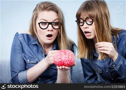 Friendship, human relations concept. Two crazy women friends or sisters wearing jeans shirts and eyeglasses on stick, thinking about solving problem holding fake brain. Two crazy women friends thinking