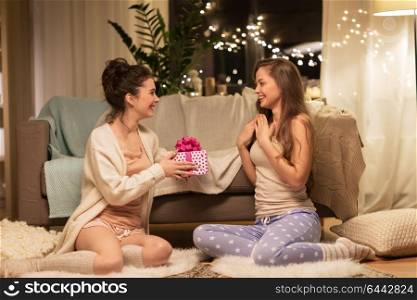 friendship, hygge and christmas concept - happy young woman or teenage girl giving present to friend at home pajama party. happy female friends with christmas gift at home