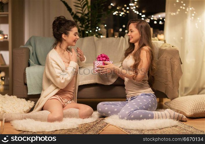 friendship, hygge and christmas concept - happy young woman or teenage girl giving present to friend at home pajama party. happy female friends with christmas gift at home. happy female friends with christmas gift at home