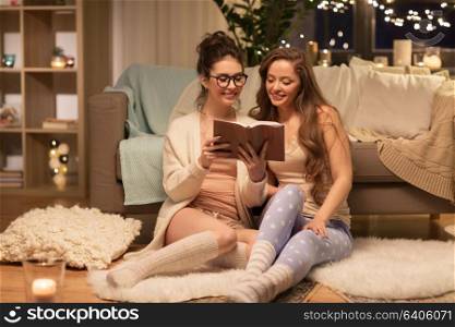 friendship, hygge and pajama party concept - happy female friends or teenage girls reading book at home. happy female friends reading book at home