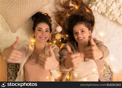 friendship, hygge and pajama party concept - happy female friends or teenage girls lying on floor and showing thumbs up at home. happy female friends showing thumbs up at home