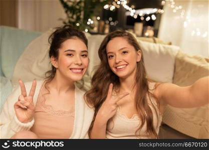 friendship, hygge and pajama party concept - happy female friends or teenage girls taking selfie at home and showing peace hand sign. female friends taking selfie at home. female friends taking selfie at home