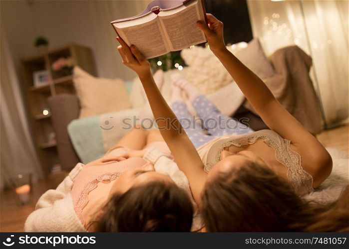 friendship, hygge and pajama party concept - happy female friends or teenage girls lying on floor and reading book at home. happy female friends reading book at home
